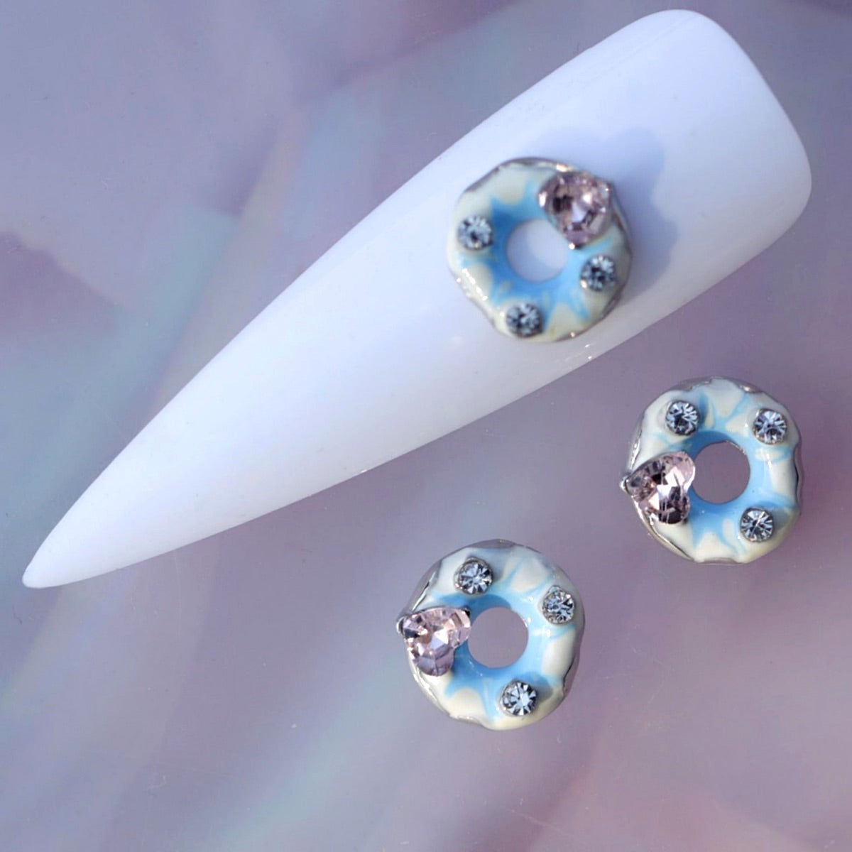 Iced glazed donut nail charm featuring pale blue and white icing, a central opening, clear rhinestone accents and a faceted pink crystal heart. This cute dessert-themed nail decoration is ideal for kawaii manicures, pastel nail art, birthday nails, party looks and playful accent designs.