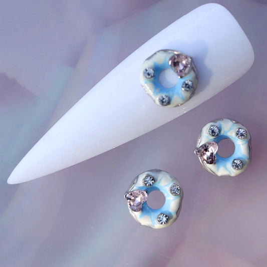 Iced glazed donut nail charm featuring pale blue and white icing, a central opening, clear rhinestone accents and a faceted pink crystal heart. This cute dessert-themed nail decoration is ideal for kawaii manicures, pastel nail art, birthday nails, party looks and playful accent designs.