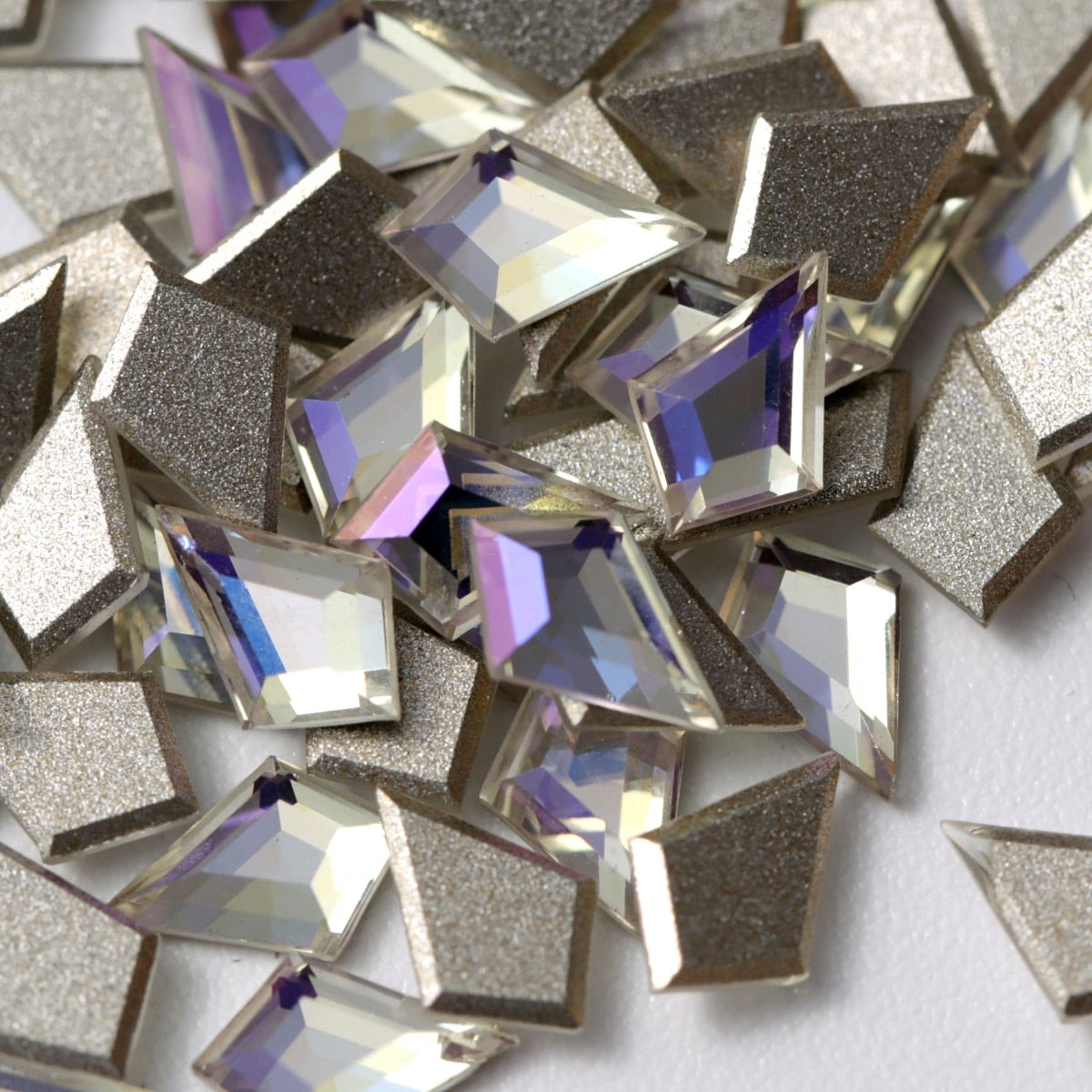 A close-up view of K9 Crystal Flatback Deltoid – Crystal Moonlight rhinestones arranged on a white surface. The angular deltoid crystals feature a clear, luminous base with cool silver, icy blue, and soft violet reflections. Their precision-cut facets and metallic silver-foil backing create a sophisticated, multidimensional sparkle, ideal for luxury nail art, bridal designs, jewelry embellishment, and high-end craft projects.