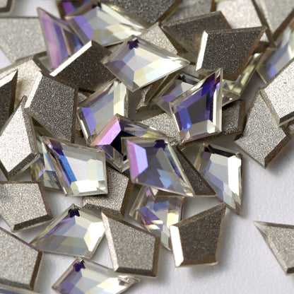 A close-up view of K9 Crystal Flatback Deltoid – Crystal Moonlight rhinestones arranged on a white surface. The angular deltoid crystals feature a clear, luminous base with cool silver, icy blue, and soft violet reflections. Their precision-cut facets and metallic silver-foil backing create a sophisticated, multidimensional sparkle, ideal for luxury nail art, bridal designs, jewelry embellishment, and high-end craft projects.