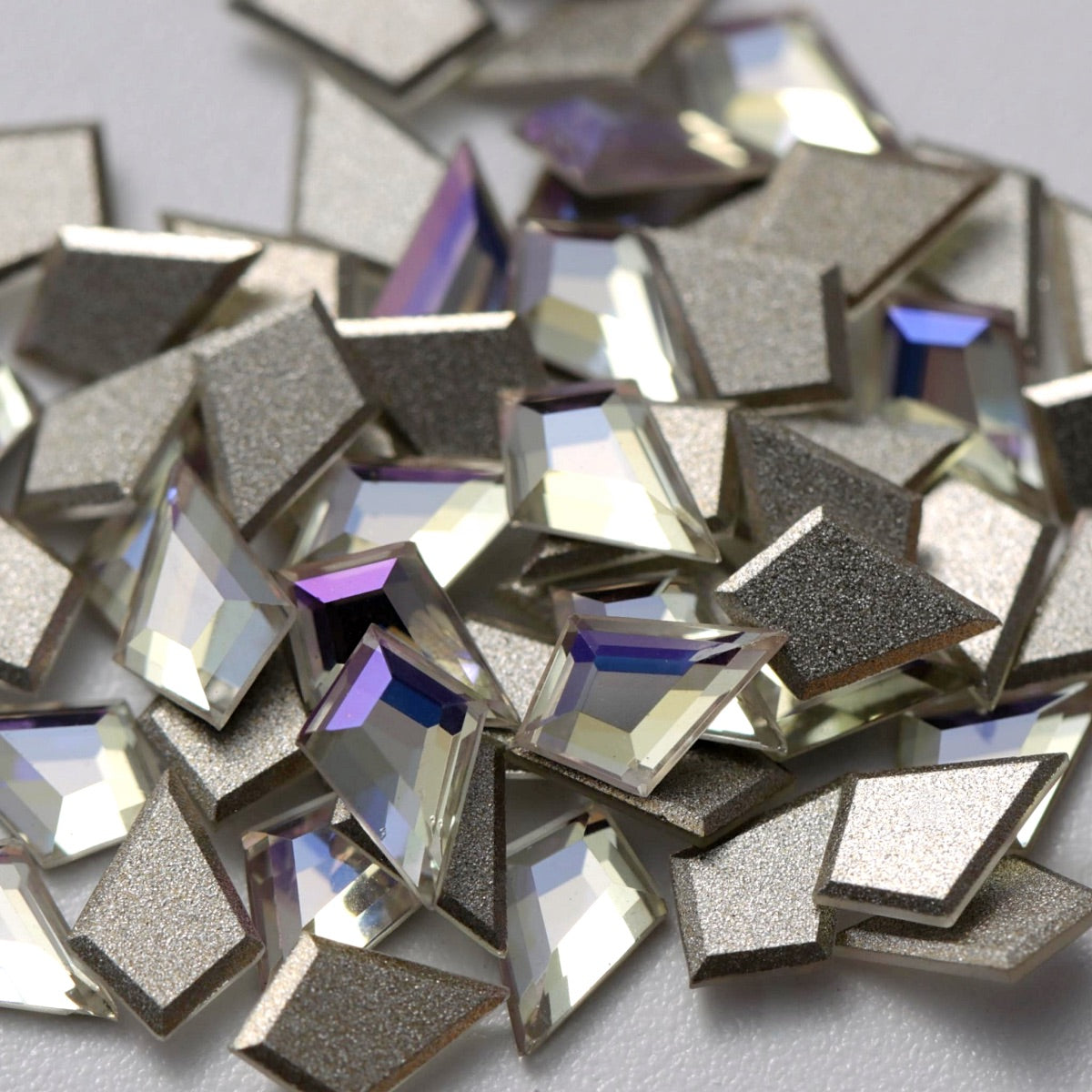 A detailed arrangement of Crystal Moonlight K9 flatback deltoid rhinestones displayed on a white background. The geometric crystals reflect cool white, pale blue, silver, and subtle lavender highlights, while the silver-foil backing enhances their brilliance and depth. Perfect for creating elegant geometric nail designs, bridal details, statement jewelry, fashion accessories, and refined DIY embellishments.