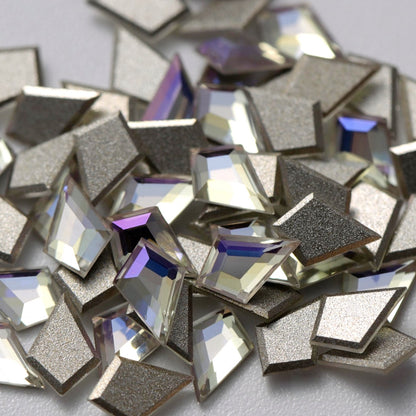 A detailed arrangement of Crystal Moonlight K9 flatback deltoid rhinestones displayed on a white background. The geometric crystals reflect cool white, pale blue, silver, and subtle lavender highlights, while the silver-foil backing enhances their brilliance and depth. Perfect for creating elegant geometric nail designs, bridal details, statement jewelry, fashion accessories, and refined DIY embellishments.