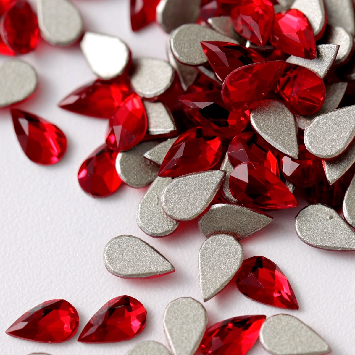 A close-up view of vibrant Siam-red K9 Crystal Flatback Drop rhinestones arranged on a white surface. The teardrop-shaped crystals feature precision-cut facets, intense ruby-red color, and reflective metallic silver backing, creating luxurious multidimensional sparkle. Ideal for bold nail art, bridal details, jewelry embellishment, fashion accessories, and elegant crafts.