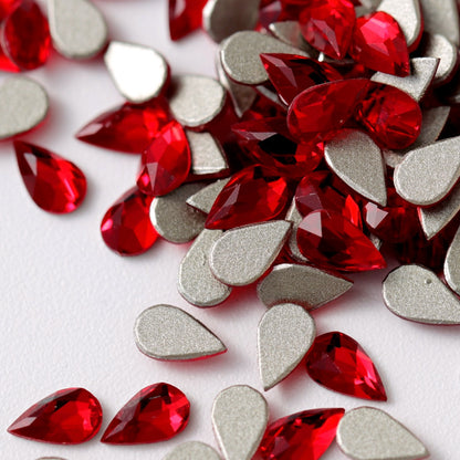 A close-up view of vibrant Siam-red K9 Crystal Flatback Drop rhinestones arranged on a white surface. The teardrop-shaped crystals feature precision-cut facets, intense ruby-red color, and reflective metallic silver backing, creating luxurious multidimensional sparkle. Ideal for bold nail art, bridal details, jewelry embellishment, fashion accessories, and elegant crafts.