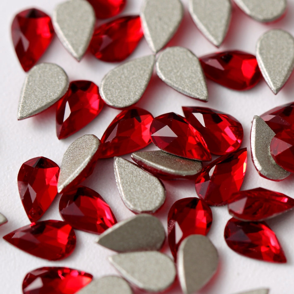 A detailed arrangement of Siam-red K9 Crystal Flatback Drop rhinestones displayed on a white background. Their elegant teardrop shape, brilliant red color, faceted surfaces, and silver foil backing create dramatic light reflection and refined depth. Perfect for statement nail designs, bridal accents, jewelry, fashion accessories, and creative embellishments.
