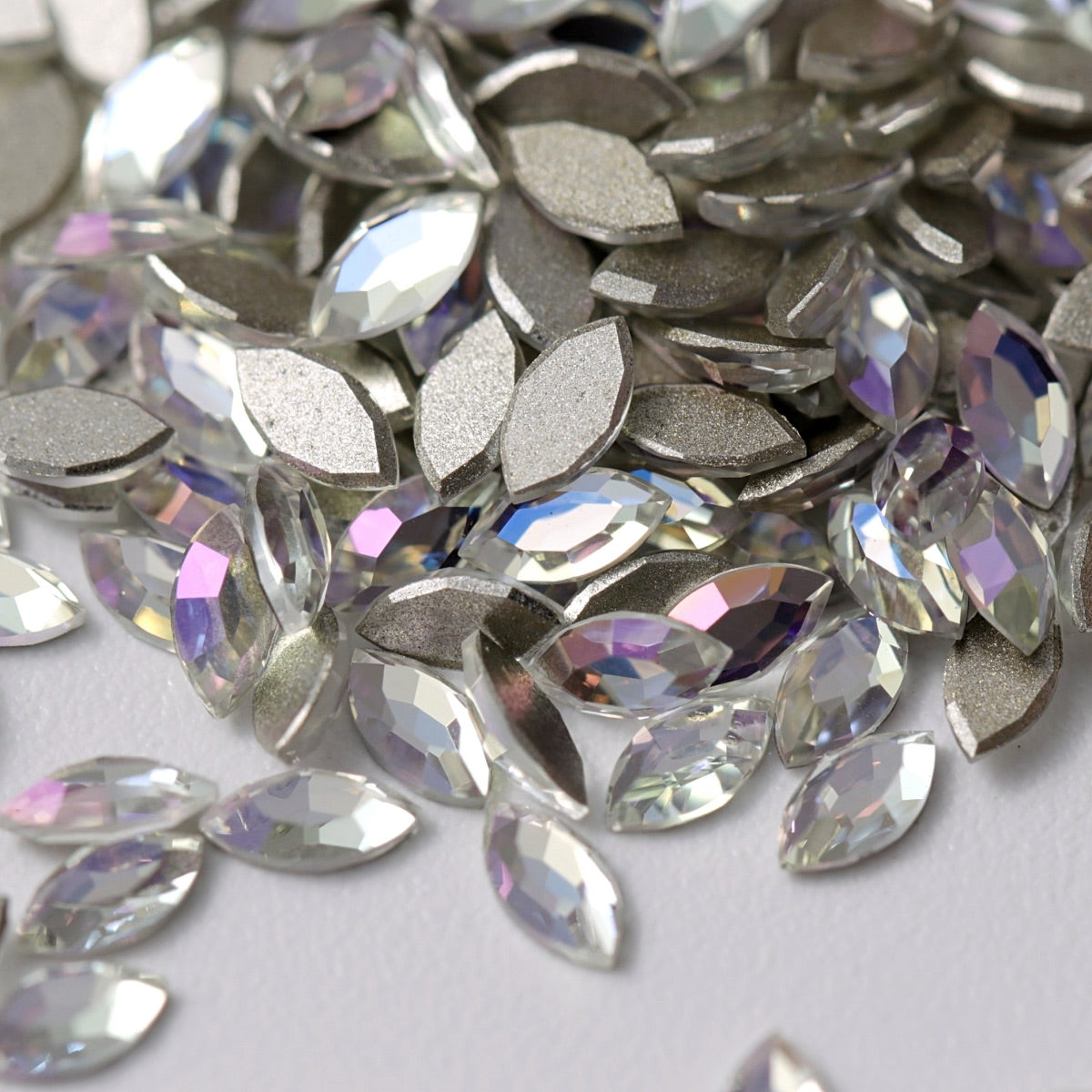 A close-up view of Crystal Moonlight K9 flatback Navette rhinestones arranged on a white surface. The elongated crystals feature a clear, luminous finish with delicate reflections of icy blue, soft violet, and pale silver. Their faceted surfaces and metallic silver-foil backing create refined, multidimensional sparkle—perfect for luxury nail art, bridal details, jewelry, and elegant crafts.