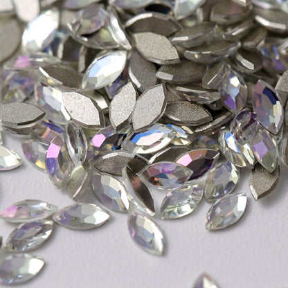 A close-up view of Crystal Moonlight K9 flatback Navette rhinestones arranged on a white surface. The elongated crystals feature a clear, luminous finish with delicate reflections of icy blue, soft violet, and pale silver. Their faceted surfaces and metallic silver-foil backing create refined, multidimensional sparkle—perfect for luxury nail art, bridal details, jewelry, and elegant crafts.