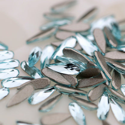 Close-up of scattered faceted K9 Crystal Flatback Raindrop Aquamarine Crystal 6×2mm with silver glitter edges on a light beige background.
