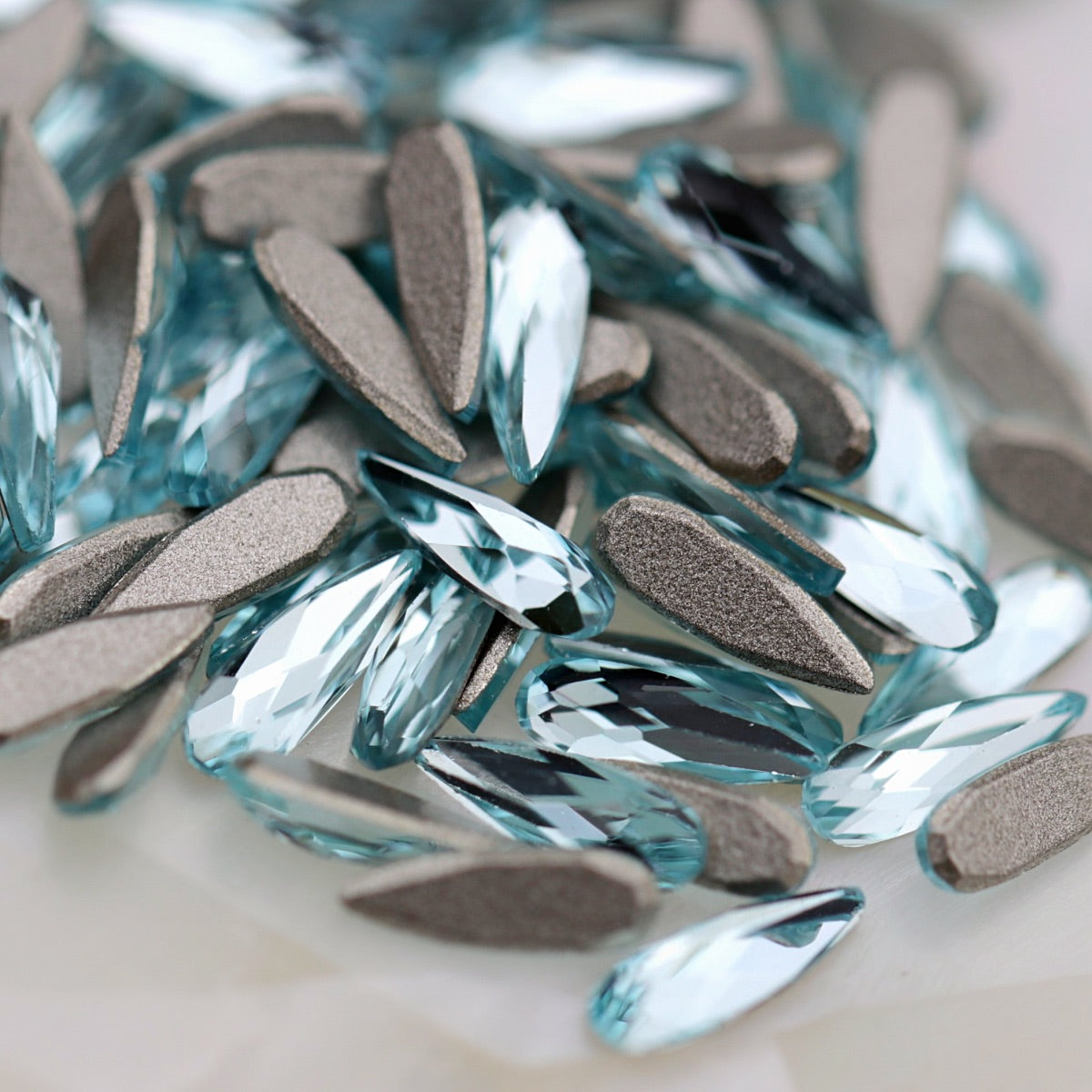 A pile of K9 Crystal Flatback Raindrop Aquamarine Crystal 6×2mm rhinestones with textured silver backing plates, photographed close-up on a light neutral surface.