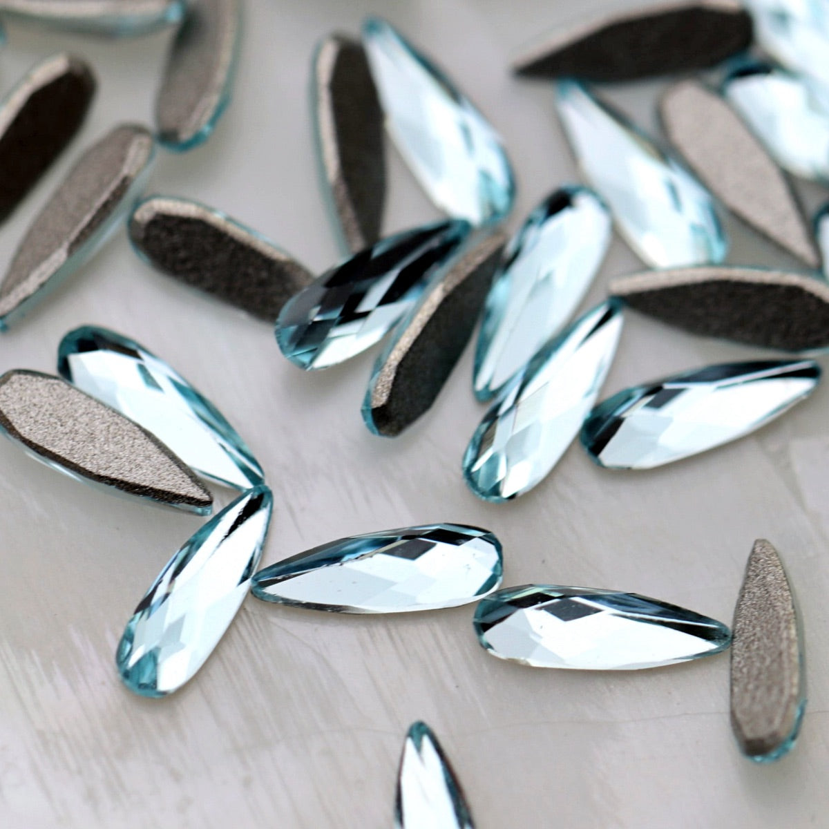 K9 Crystal Flatback Raindrop in Aquamarine Crystal, measuring 6×2mm. The elongated teardrop-shaped crystal has a narrow pointed tip, rounded lower end and faceted front surface. Its transparent light-blue colour reflects pale aqua, icy blue, turquoise, seafoam, soft teal and silvery tones. Ideal for bridal nails, ocean-inspired nail art, mermaid designs, floral manicures, crystal clusters and elegant nail decorations.