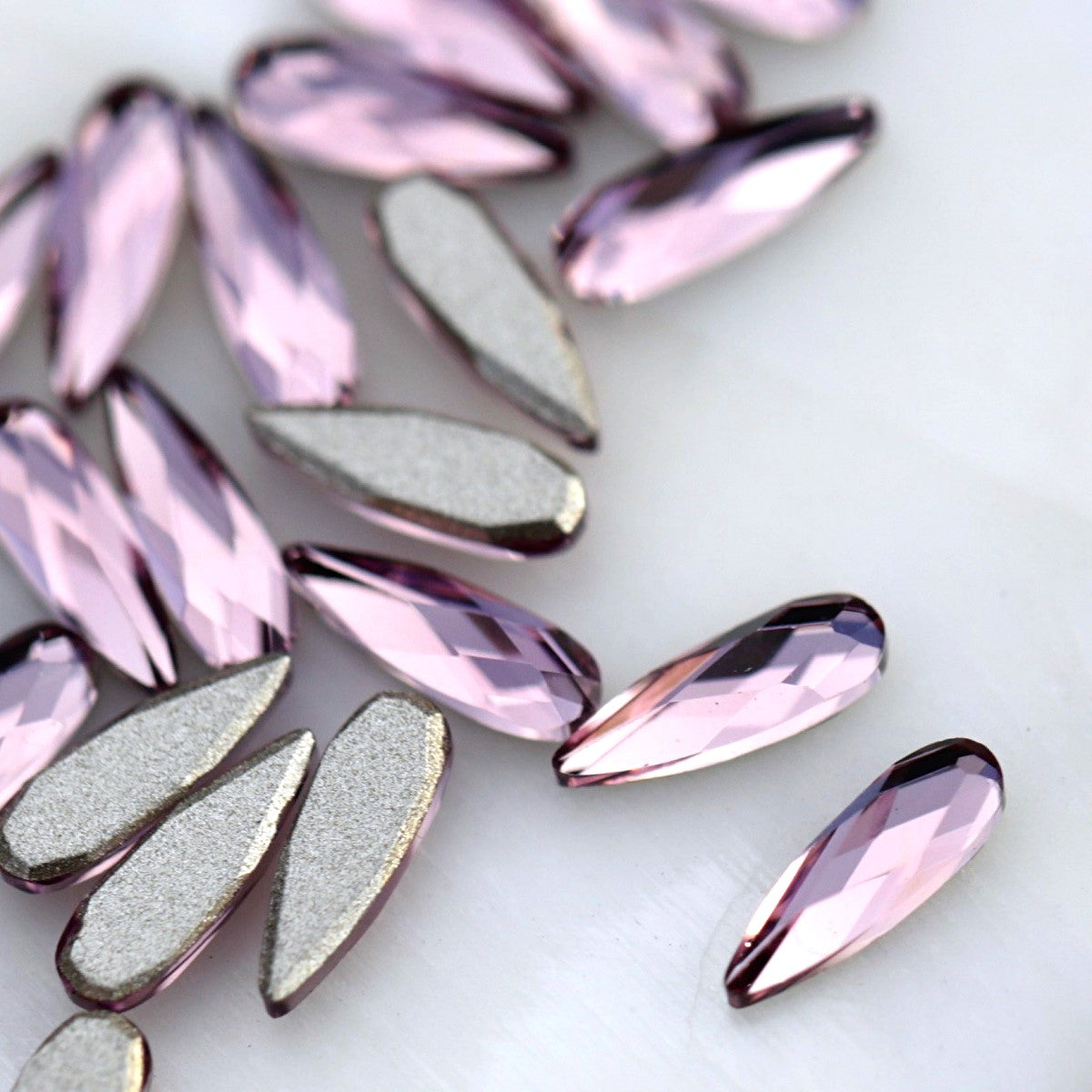 Close-up of slim 6×2mm K9 Crystal raindrops in Light Amethyst, showing faceted pale lavender surfaces with soft lilac and pink reflections.