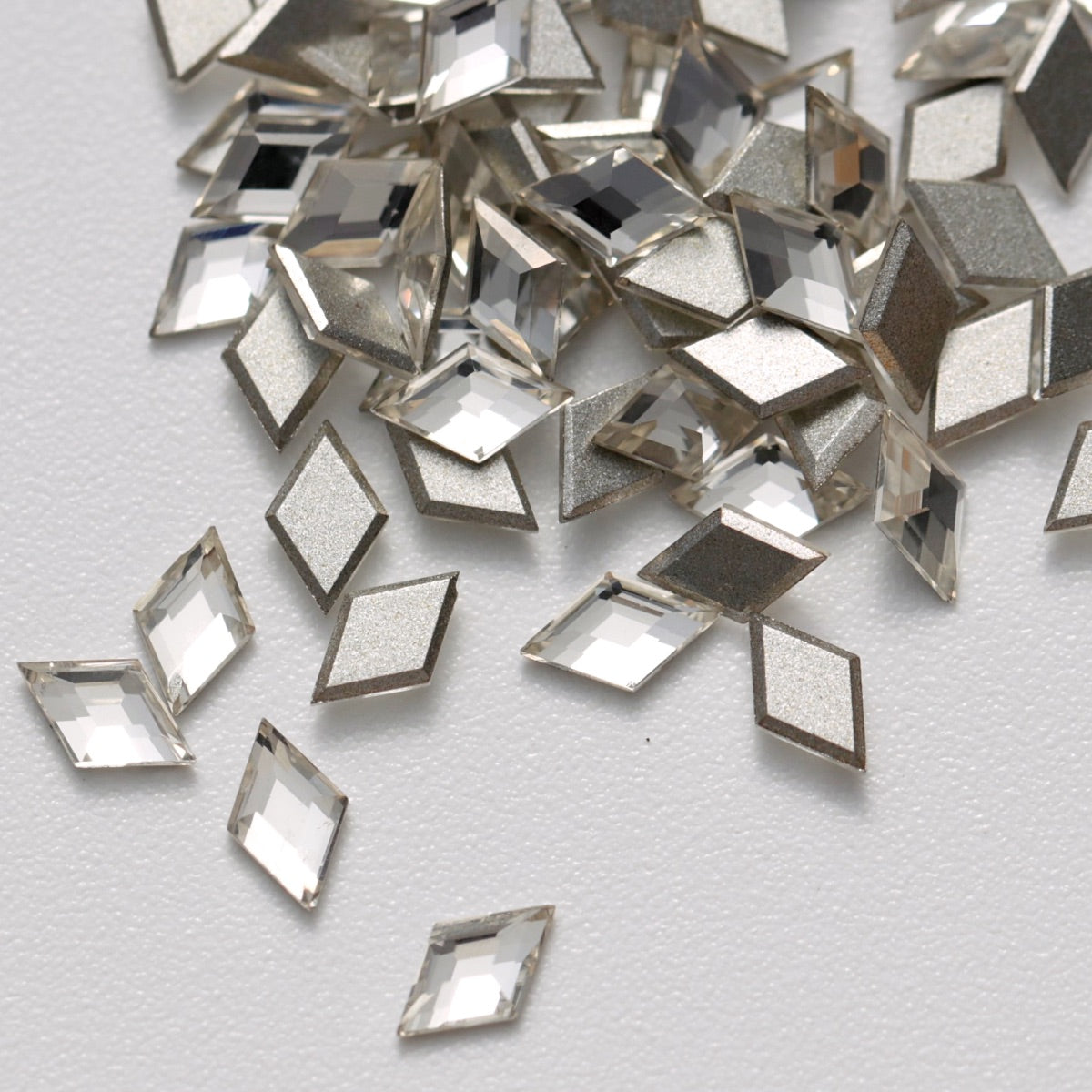 A close-up view of clear Crystal K9 flatback rhombus stones arranged on a white surface. The diamond-shaped crystals feature precision-cut facets and reflective silver foil backings that create bright, multidimensional sparkle. Ideal for geometric nail art, jewelry decoration, fashion accessories, and creative DIY projects.