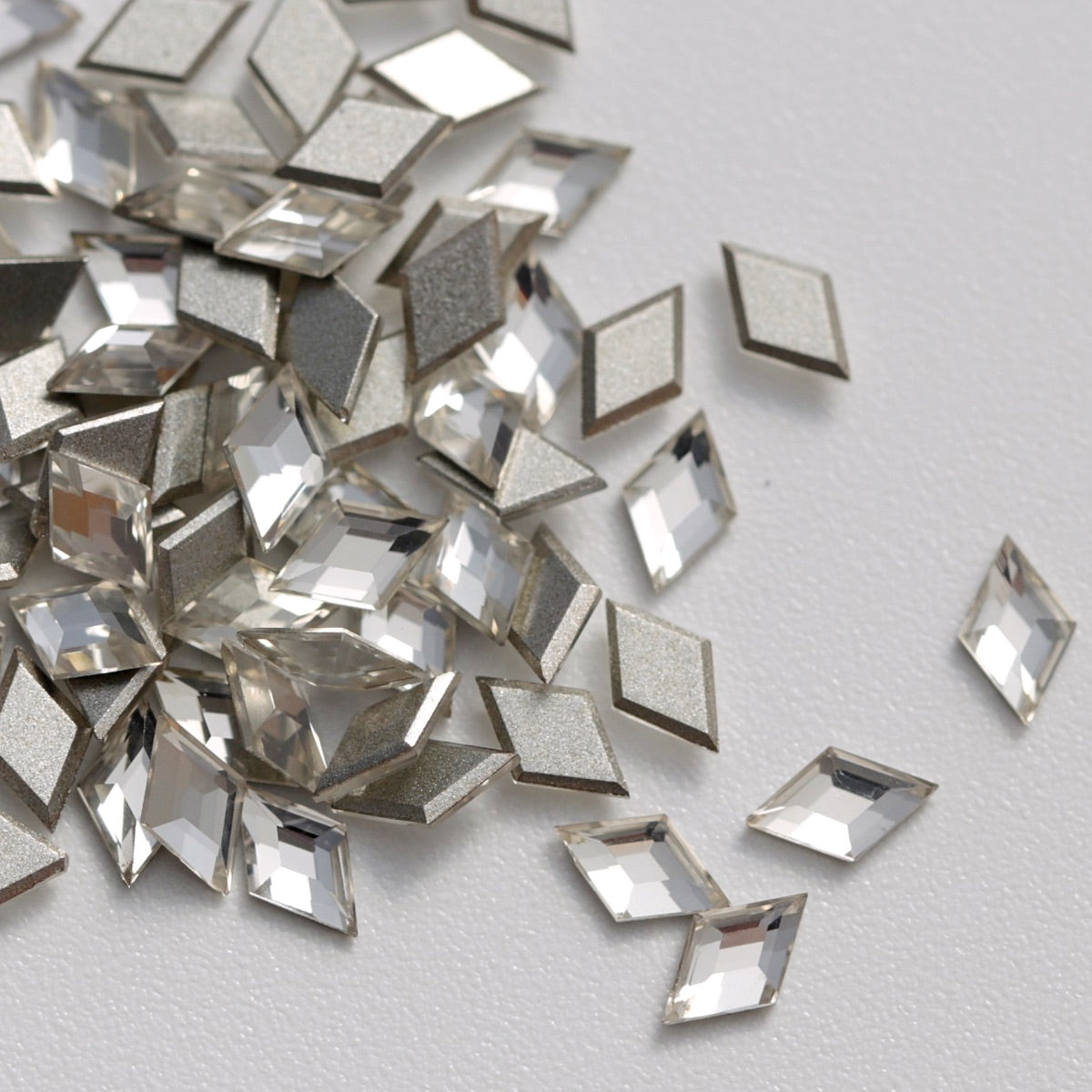 A detailed close-up of clear K9 crystal flatback rhombus stones displayed on a white background. Their transparent finish, sharp geometric shape, and reflective silver backings enhance light reflection, making them suitable for elegant nail designs, jewelry accents, fashion embellishments, and creative crafts.