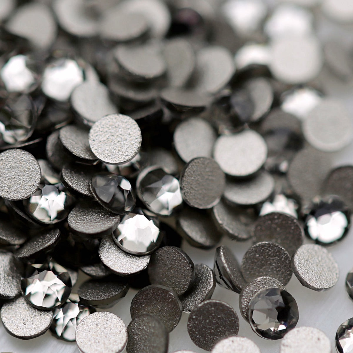 Black Diamond K9 flatback rhinestones displayed from the front and back, highlighting their reflective dark-grey faceted tops and flat backs for nail-art application.