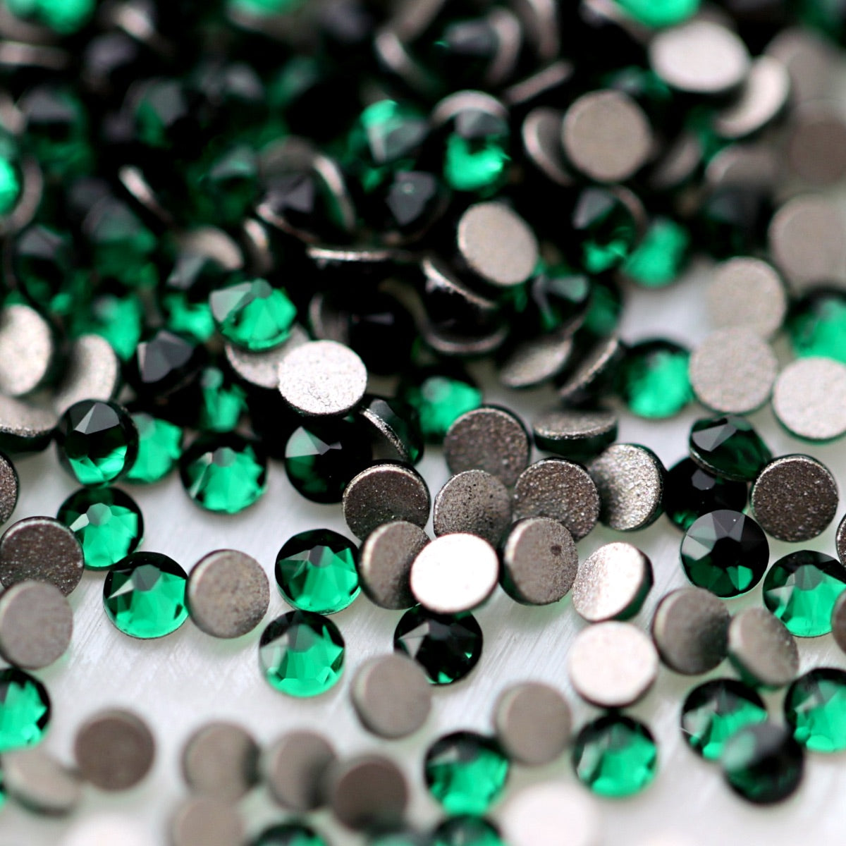 Close-up of round K9 flatback rhinestones in deep Emerald green, showing defined faceting, vivid green reflections and flat backs for nail-art application.