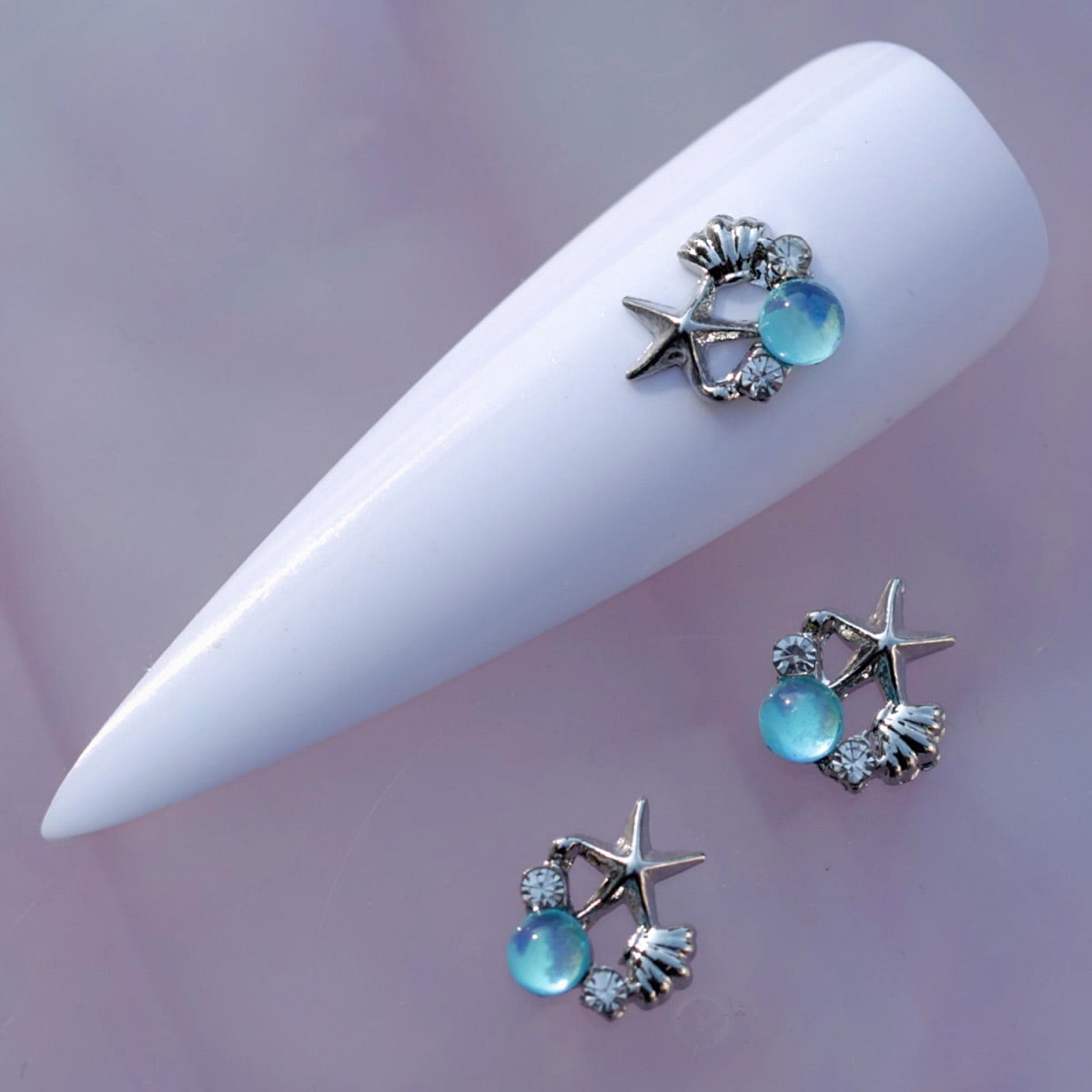Sea-themed nail charm featuring a silver-tone starfish, shell, clear rhinestones and a translucent aquamarine blue glass cabochon. This ocean-inspired decoration is ideal for summer nail art, beach manicures, mermaid designs and aqua accent nails.