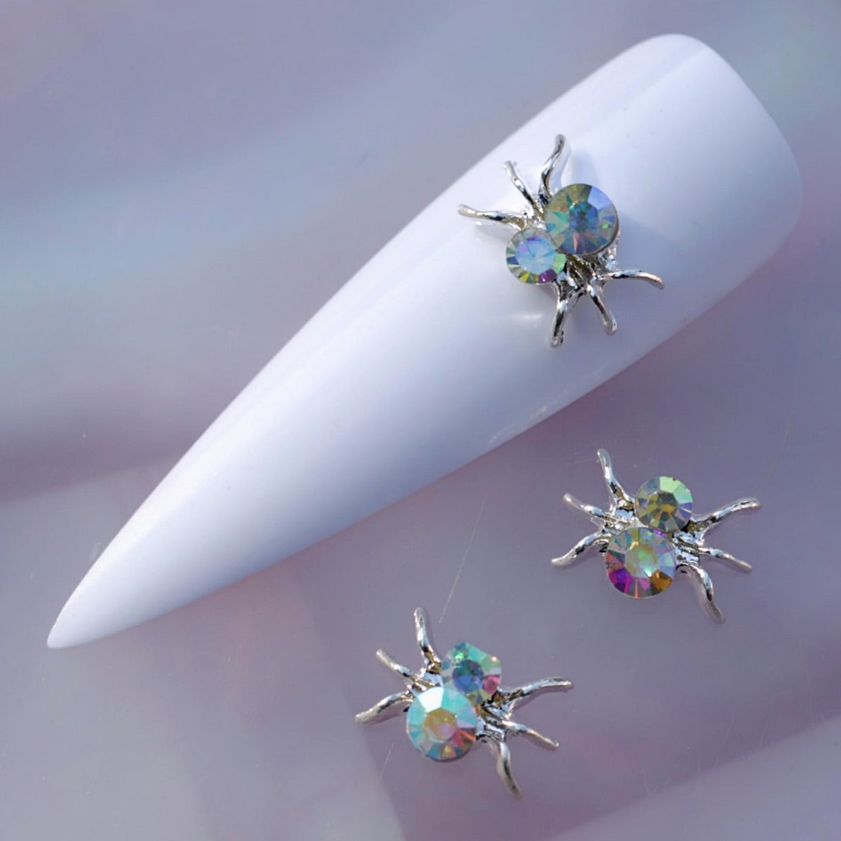 Silver-tone spider nail charm featuring delicate curved legs and two faceted iridescent crystal rhinestones. The crystals reflect blue, green, purple, pink, yellow and silvery tones, making this sparkling spider decoration ideal for Halloween manicures, gothic nail art, spiderweb designs and creative accent nails.