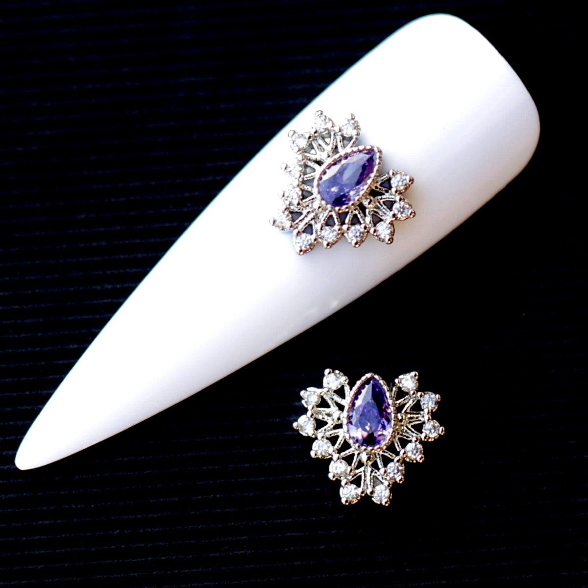 Decorative nail charm featuring a central faceted amethyst-coloured teardrop cubic zirconia crystal surrounded by clear crystal accents and a silver-tone setting. The jewellery-inspired nail decoration has a rich purple, violet and lavender sparkle and is suitable for bridal manicures, evening looks, glamour designs and elegant accent nail art.