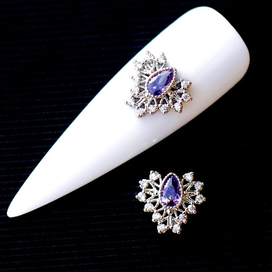 Decorative nail charm featuring a central faceted amethyst-coloured teardrop cubic zirconia crystal surrounded by clear crystal accents and a silver-tone setting. The jewellery-inspired nail decoration has a rich purple, violet and lavender sparkle and is suitable for bridal manicures, evening looks, glamour designs and elegant accent nail art.