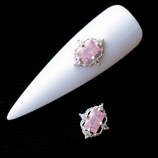 Silver-tone nail charm featuring a large faceted pink baguette-shaped cubic zirconia crystal in the centre, surrounded by silver-tone details and small clear crystal accents. The decorative nail jewellery piece has a soft romantic appearance and is suitable for bridal manicures, wedding nails, glamour designs and elegant accent nail art.