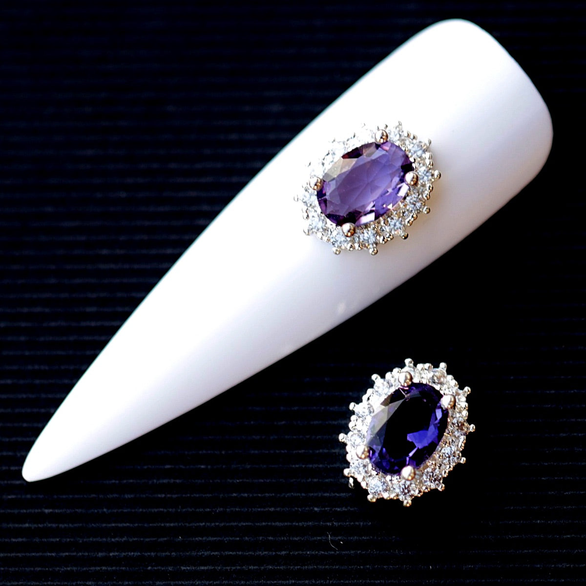 Oval nail charm featuring a faceted amethyst-coloured cubic zirconia crystal in the centre, surrounded by a halo of clear decorative crystals and a silver-tone setting. The jewellery-inspired nail decoration has rich violet, purple, lavender and deep plum reflections and is suitable for bridal manicures, evening looks, glamour designs and elegant accent nails.