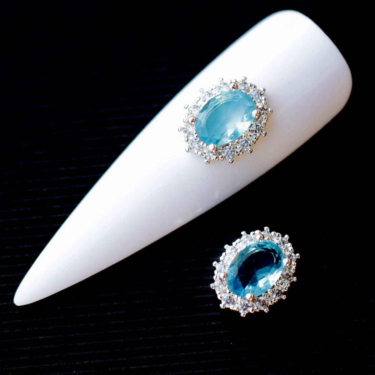 Oval nail charm featuring a faceted aquamarine cubic zirconia crystal in the centre, surrounded by a halo of clear decorative crystals and a silver-tone setting. The jewellery-inspired nail decoration has bright aqua, turquoise and cool blue reflections and is suitable for bridal manicures, ocean-themed designs, mermaid nail art, summer looks and elegant accent nails.