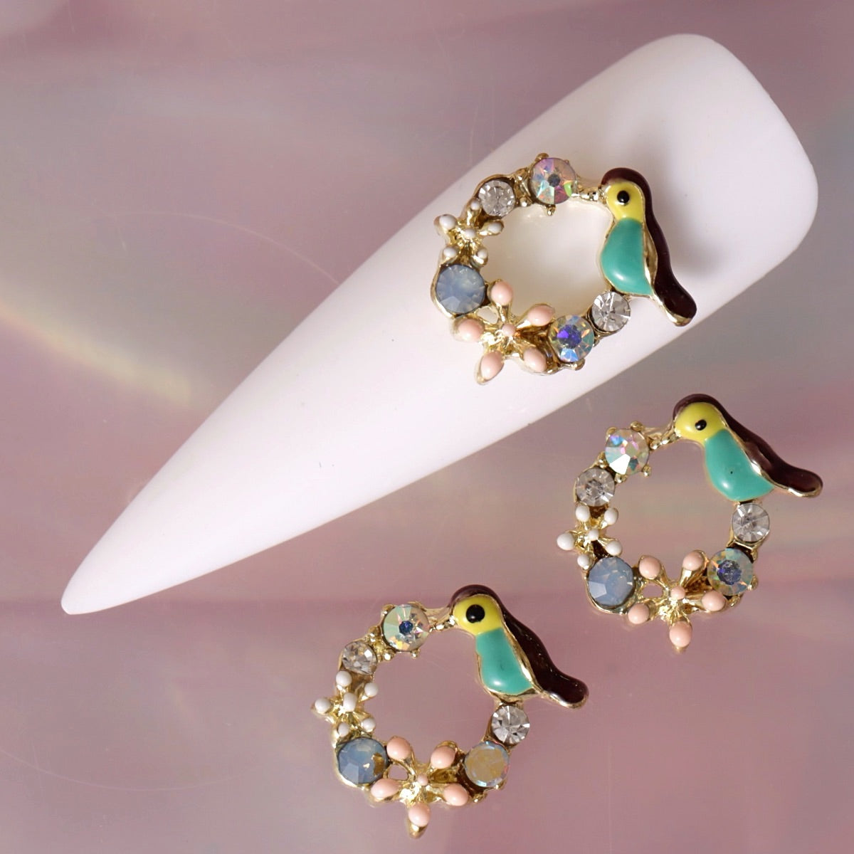 Bird composition nail charm featuring a turquoise and yellow bird, dark wing detail, sparkling crystals, soft pink and white flowers, pearls and decorative gold-tone elements. This artistic decoration is ideal for tropical, summer, garden-inspired, romantic and statement nail art.