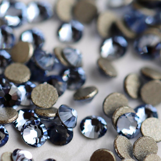 A close-up pile of Swarovski 2058 Light Sapphire flatback rhinestones in a light blue color, showing both the clear crystal tops and the textured metallic gold-toned bases on a light neutral background.