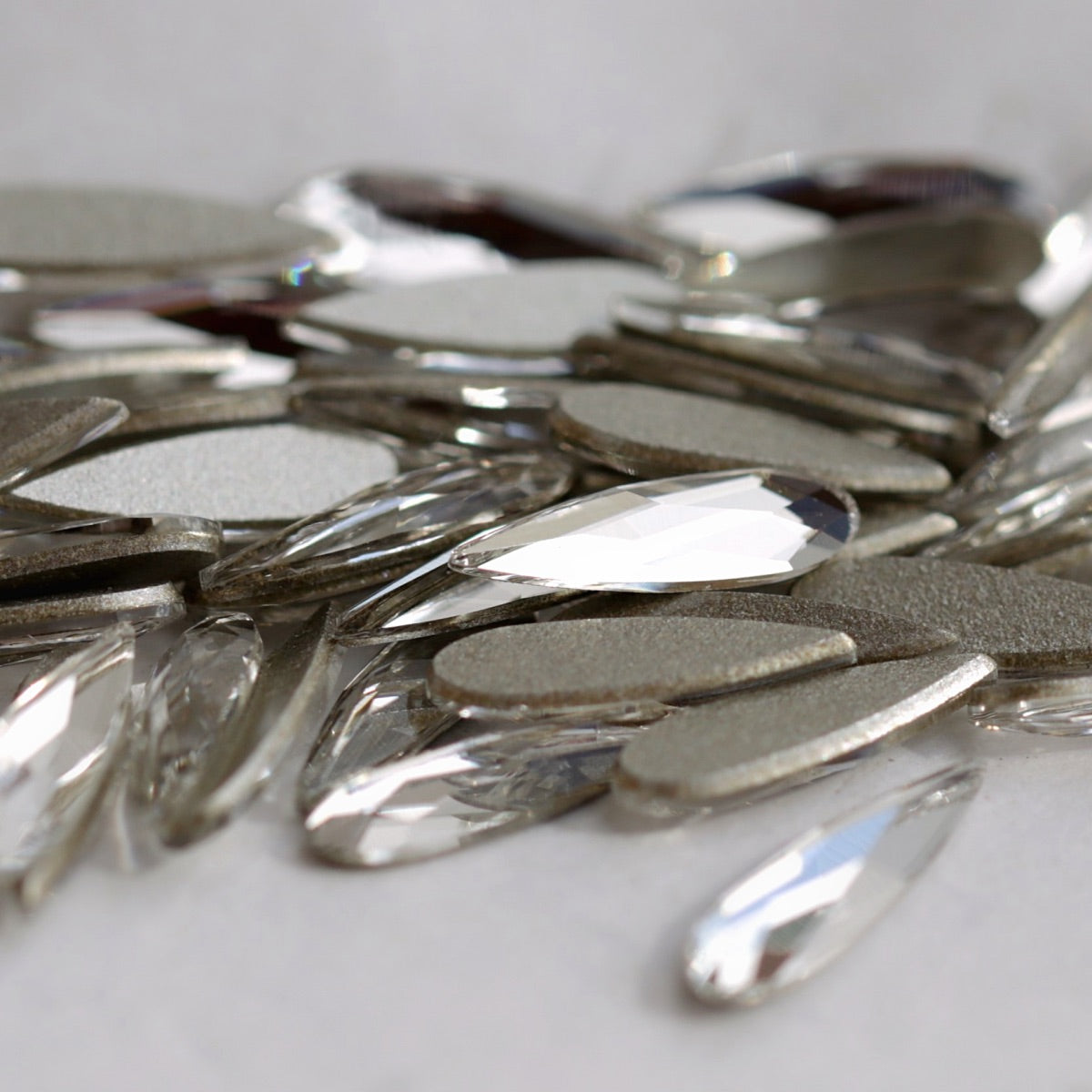 A detailed close-up of Swarovski 2304 Crystal Raindrop stones. The image highlights both the clear faceted fronts and the flat reverse sides of the long teardrop crystals, demonstrating their shape and nail-art-friendly flatback construction.