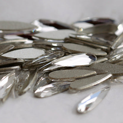 A detailed close-up of Swarovski 2304 Crystal Raindrop stones. The image highlights both the clear faceted fronts and the flat reverse sides of the long teardrop crystals, demonstrating their shape and nail-art-friendly flatback construction.
