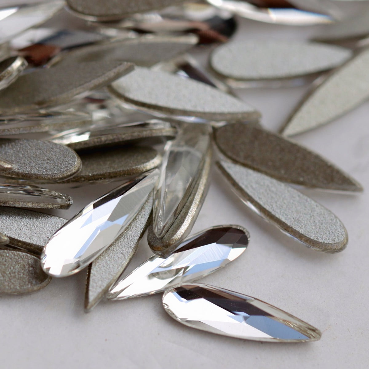 Close-up image of clear Swarovski 2304 Raindrop flatback crystals in mixed sizes. The front-facing stones show their precision-cut, reflective teardrop facets, while the reverse side shows the flat backs used for secure nail application. Ideal for detailed crystal patterns, bridal nails and elegant accent designs.