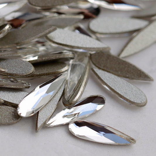 Close-up image of clear Swarovski 2304 Raindrop flatback crystals in mixed sizes. The front-facing stones show their precision-cut, reflective teardrop facets, while the reverse side shows the flat backs used for secure nail application. Ideal for detailed crystal patterns, bridal nails and elegant accent designs.