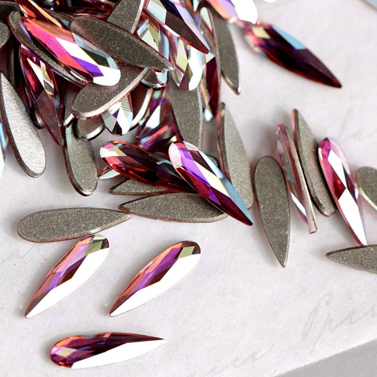 A pile of teardrop-shaped Swarovski 2304 Light Rose AB flatback rhinestones with a metallic silver backing, showing iridescent pink, purple, and blue reflections on a light gray background.