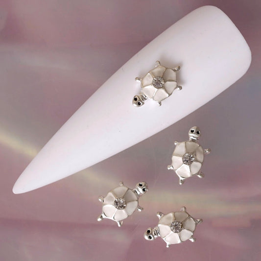 White turtle nail charm featuring a segmented white shell, polished silver-tone details, small raised limbs and a clear rhinestone in the centre. This elegant animal-inspired decoration is ideal for summer manicures, beach nail art, ocean-themed designs, bridal manicures and delicate accent nails.
